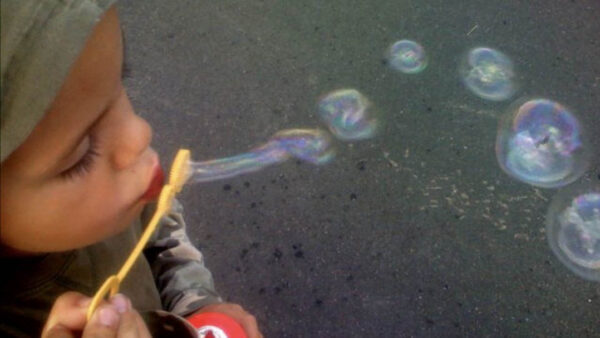 Child blowing bubbles with a yellow wand, forming soap bubbles over a dark pavement surface.