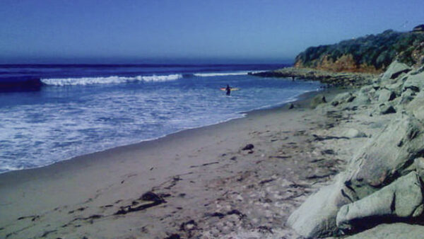 Wide sandy beach with rocky cliff on the right and gentle waves rolling in from the blue ocean; a person stands near shore in shallow water.