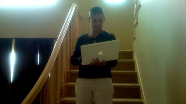 Person standing on a wooden staircase, holding an open silver MacBook and looking at the screen.