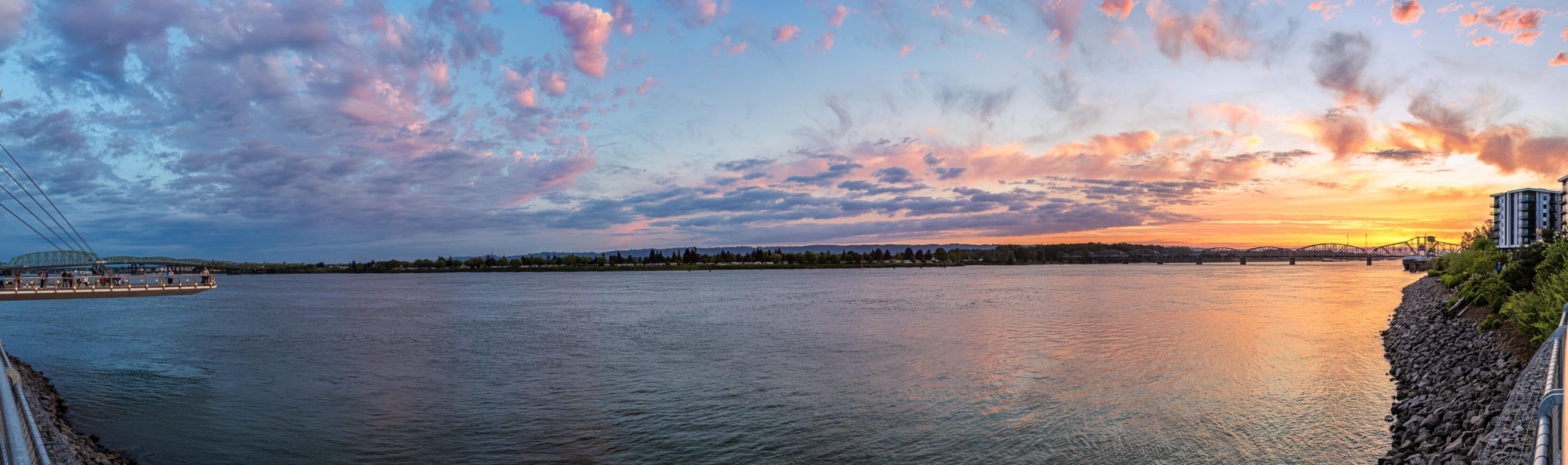 2026_05_01_Sunset_Vancouver_00023-Pano-Edit-Edit ‣ In The Viewfinder Panoramic river sunset: calm water reflecting orange-pink sky, with a pedestrian dock on the left and a distant bridge on the right.