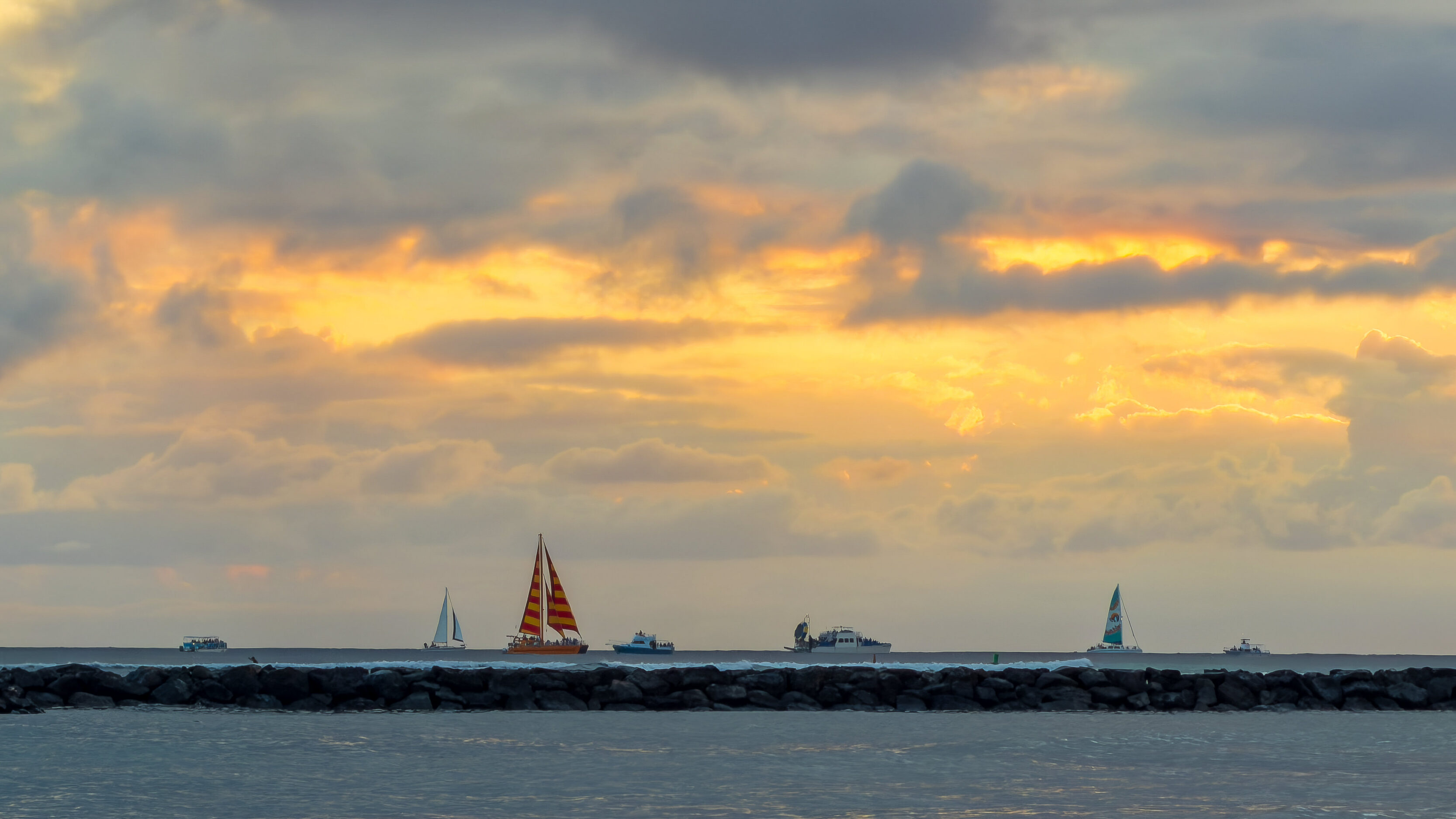 Sunset over a calm sea with several sailboats near the horizon and a rocky breakwater in the foreground. The sky glows with orange and yellow hues as clouds scatter the light.