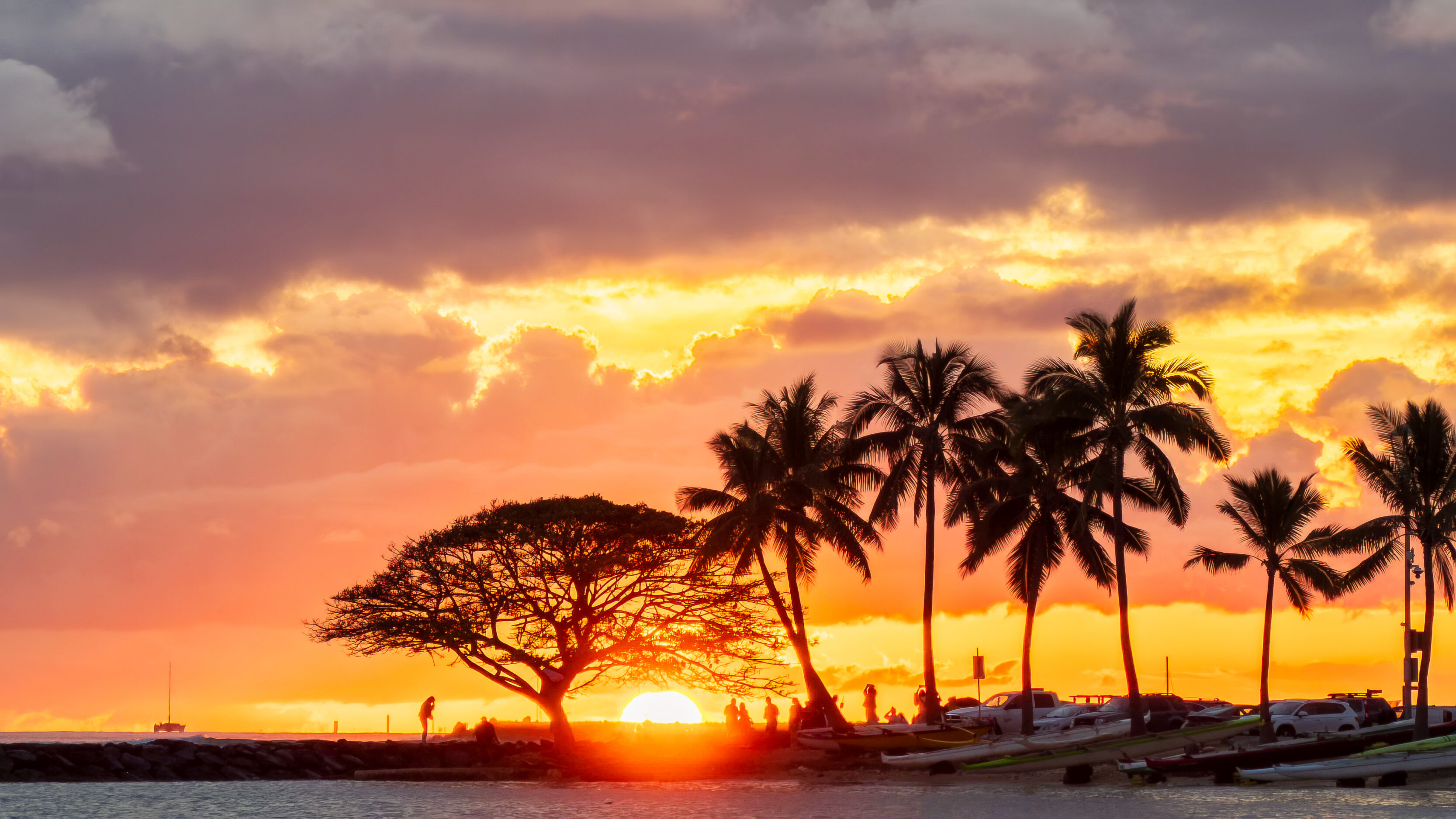 Sunset on a tropical beach with silhouetted palm trees and people along the shore.