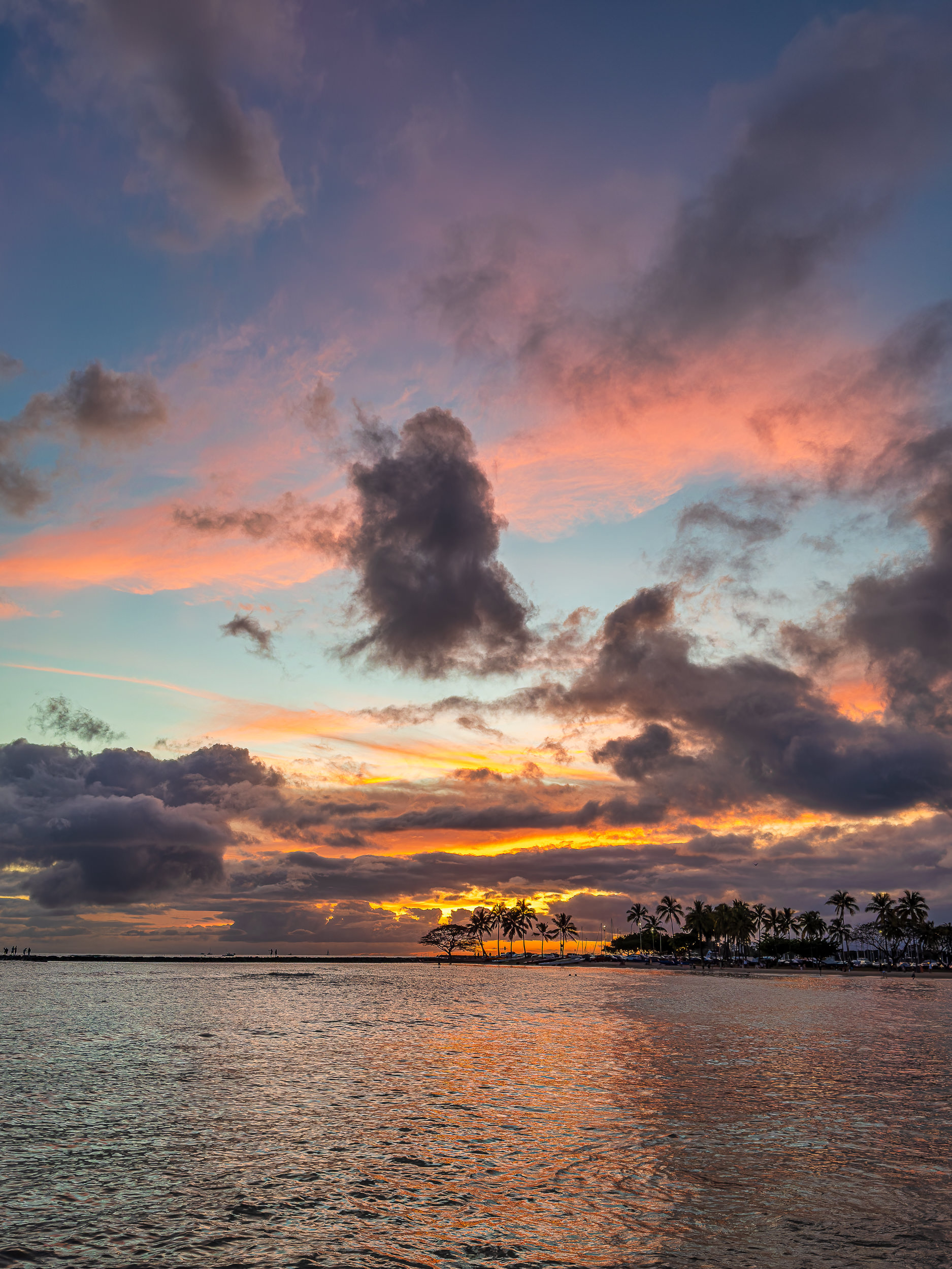 Tropical beach at sunset with orange and pink clouds over calm water and palm-tree silhouettes along the shore in the distance.