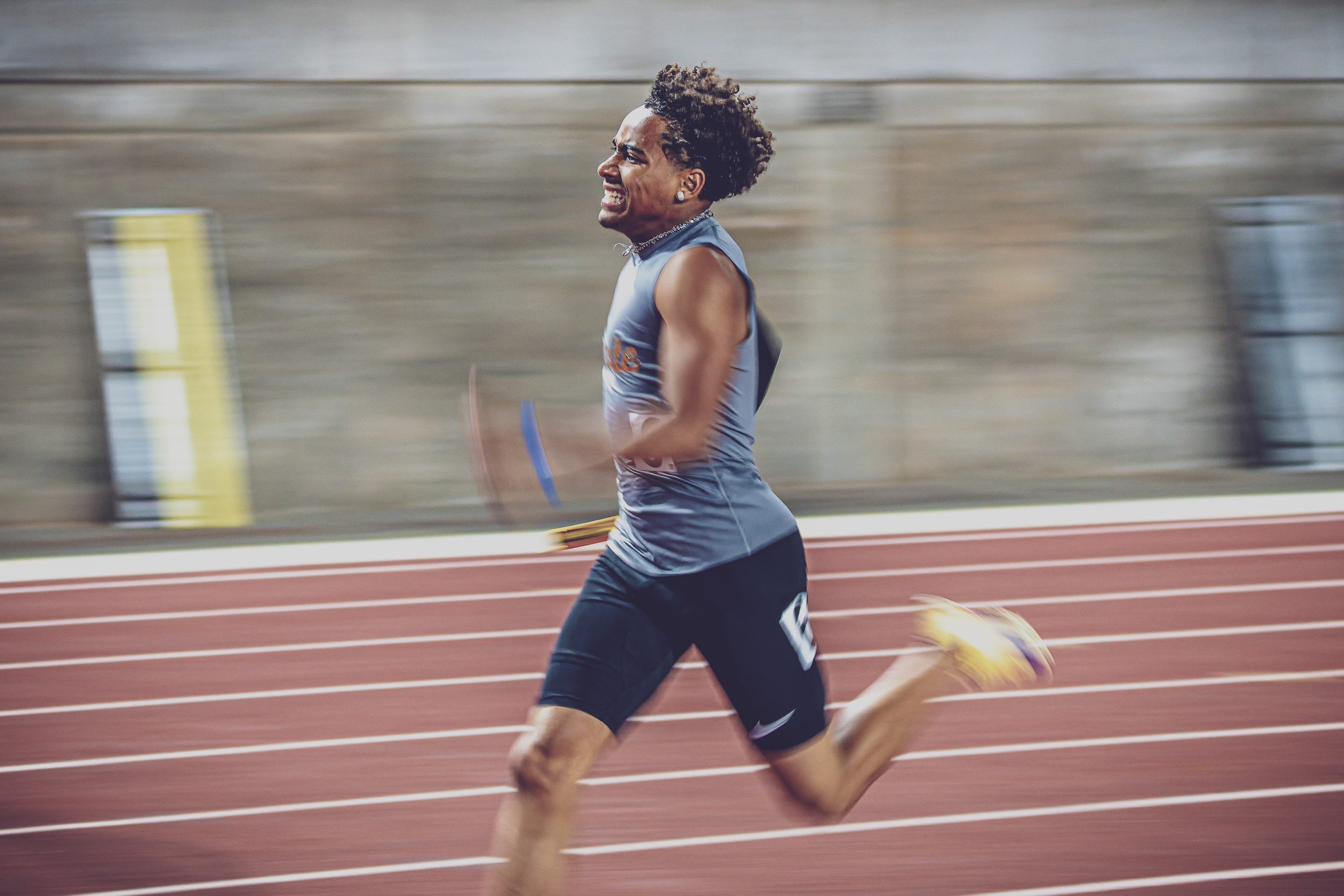 Male runner in blue athletic gear sprinting on a red track, motion blurred backdrop, determined expression.