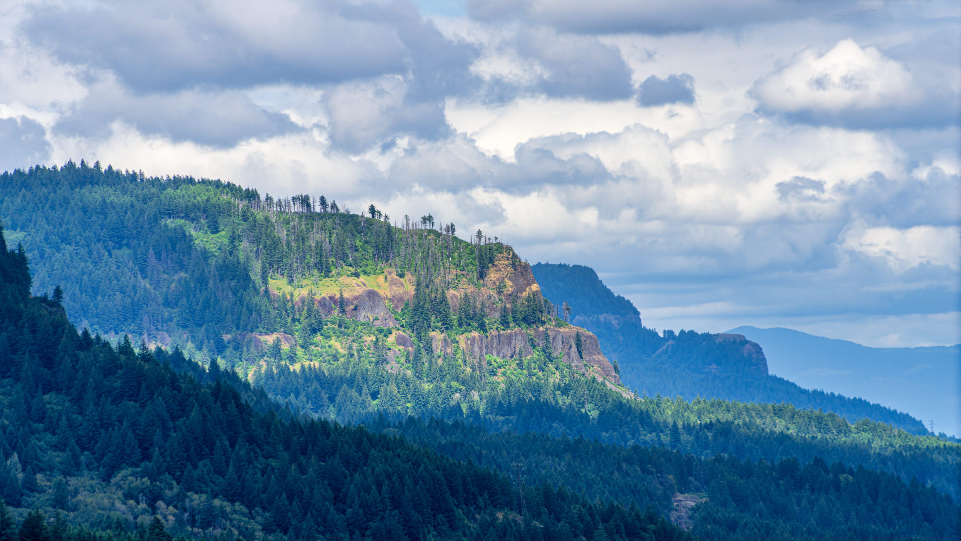 Mountain ridge with dense evergreen forest, rocky cliffs, and a cloudy sky overhead.
