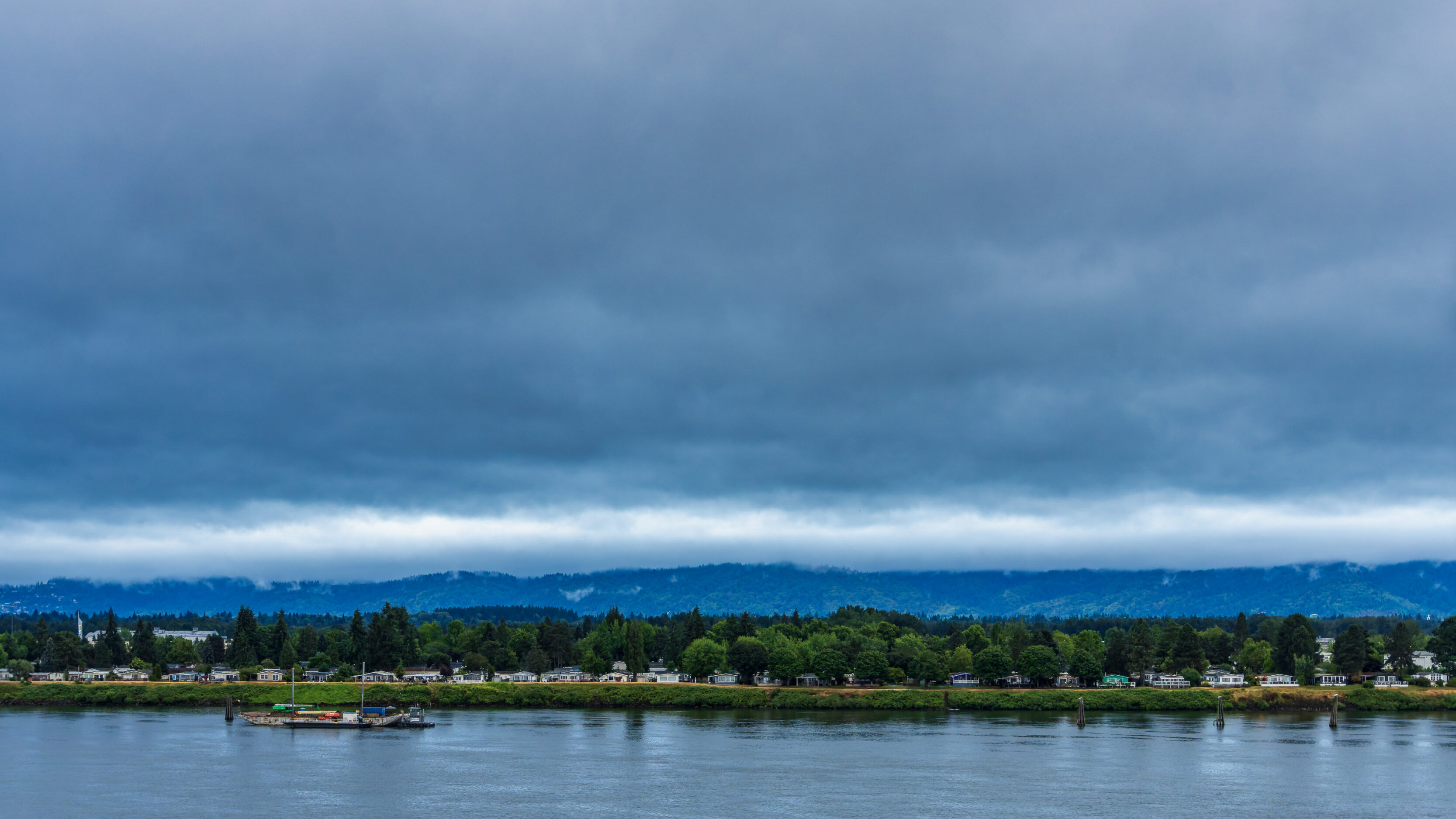 Riverside town with houses and trees along the water under a dark, stormy sky.