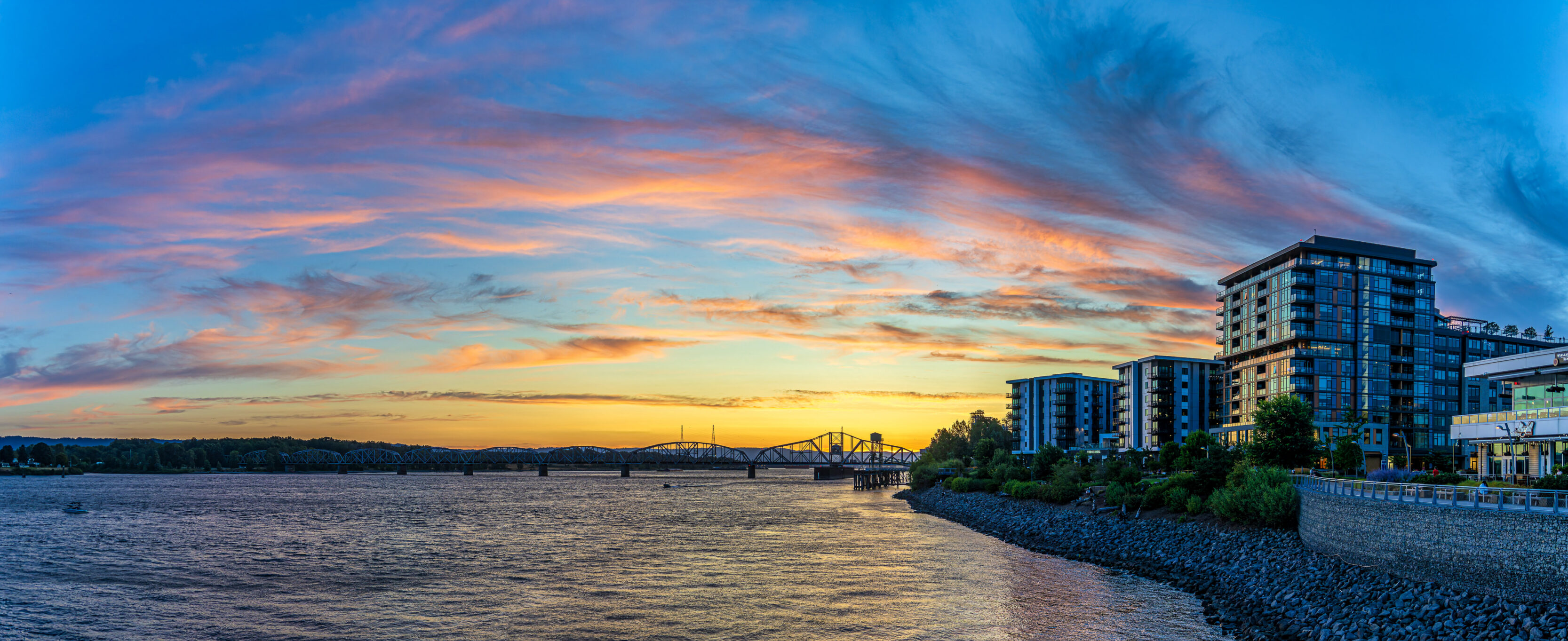 2026_07_14_Jeff_Waterfront_0042-Pano-Edit_3840 ‣ In The Viewfinder Sunset over a river with a low bridge, calm water, and modern apartment buildings along a rocky shoreline. (informative)