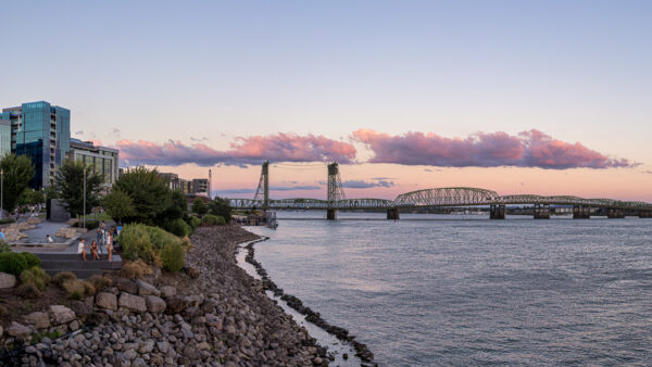 People walk along a rocky riverside promenade as modern buildings rise on the left and a steel bridge spans the calm water at sunset with pink clouds above.