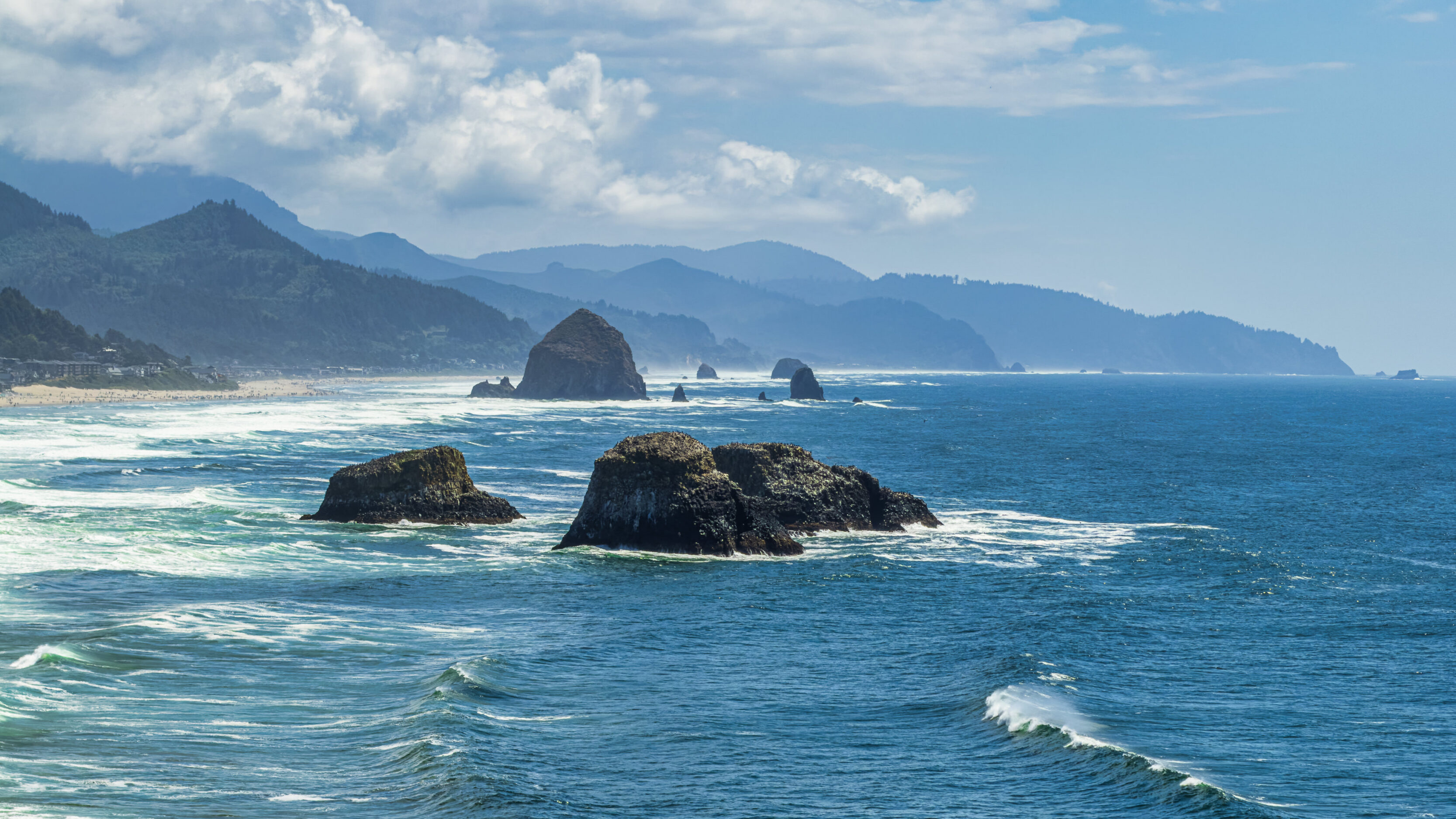 Rocky shoreline with large sea stacks rising from blue ocean, forested hills in the distance under a partly cloudy sky.