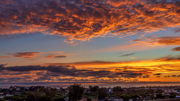 Fiery sunset over a coastal town with orange clouds lighting the sky above a calm horizon by the sea.