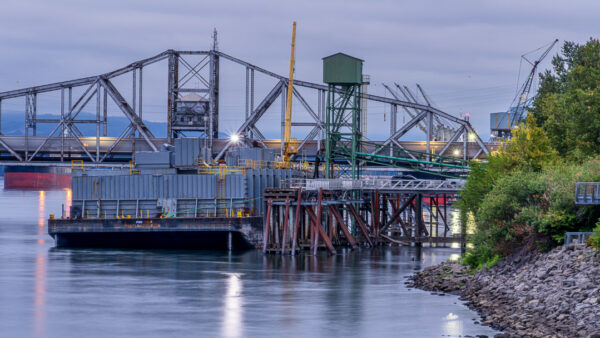 Industrial waterfront at dusk featuring a barge, crane, and lattice-truss bridge over calm water.