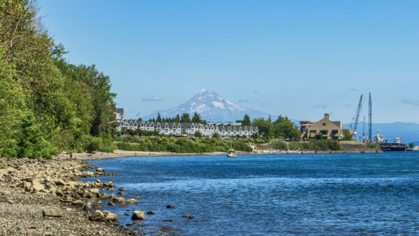 Coastal harbor with a row of white-roofed buildings along the shore, a snow‑capped mountain in the distance, and blue water in the foreground.