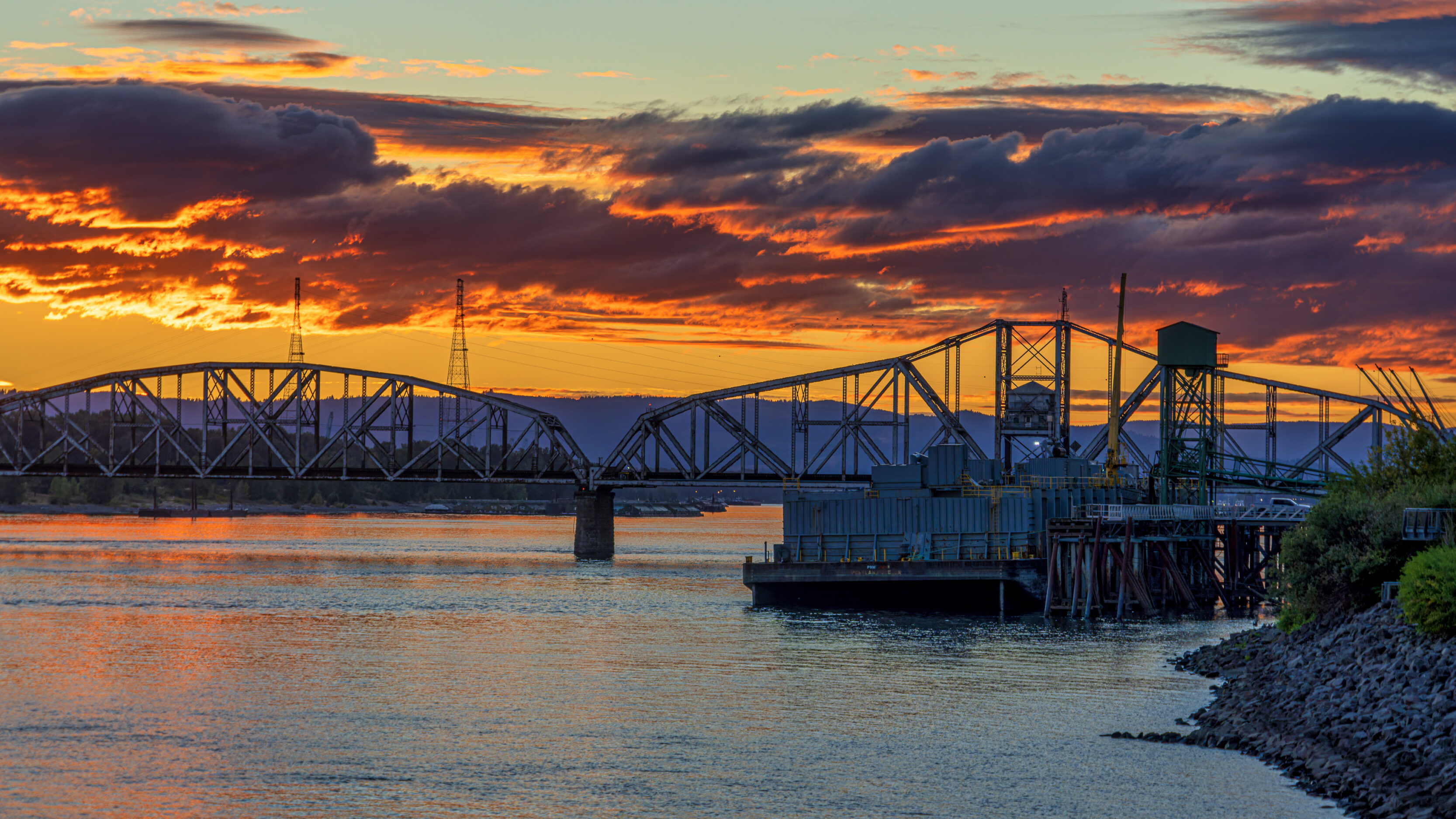 2026_08_26_Waterfront_Sunset_0038-Edit_3840 ‣ In The Viewfinder Sunset over a river with a large metal truss bridge and industrial structures on the right, orange and purple clouds lighting the sky.