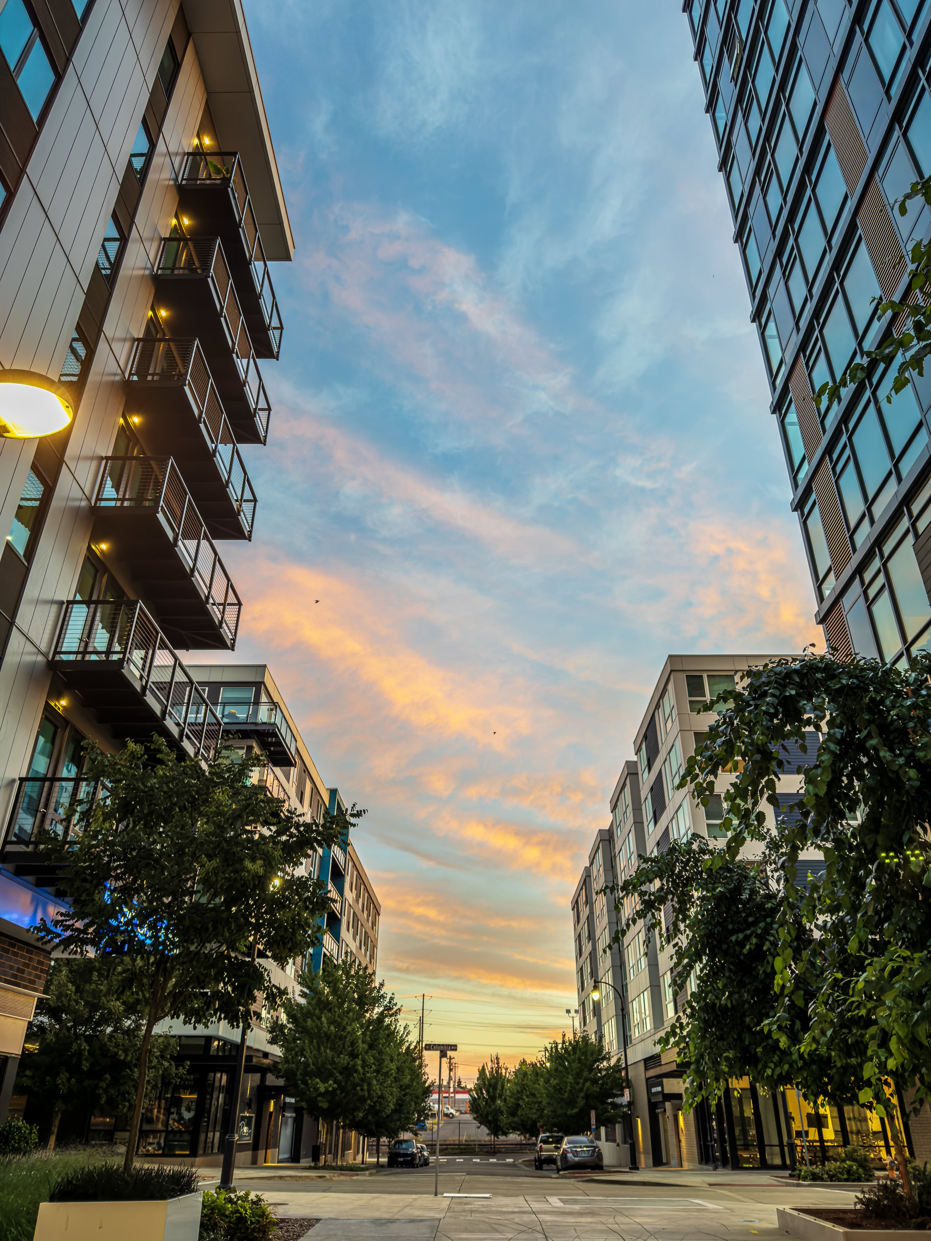 Urban street framed by modern buildings with balconies on the left and glassy façades on the right, a sunset sky overhead.