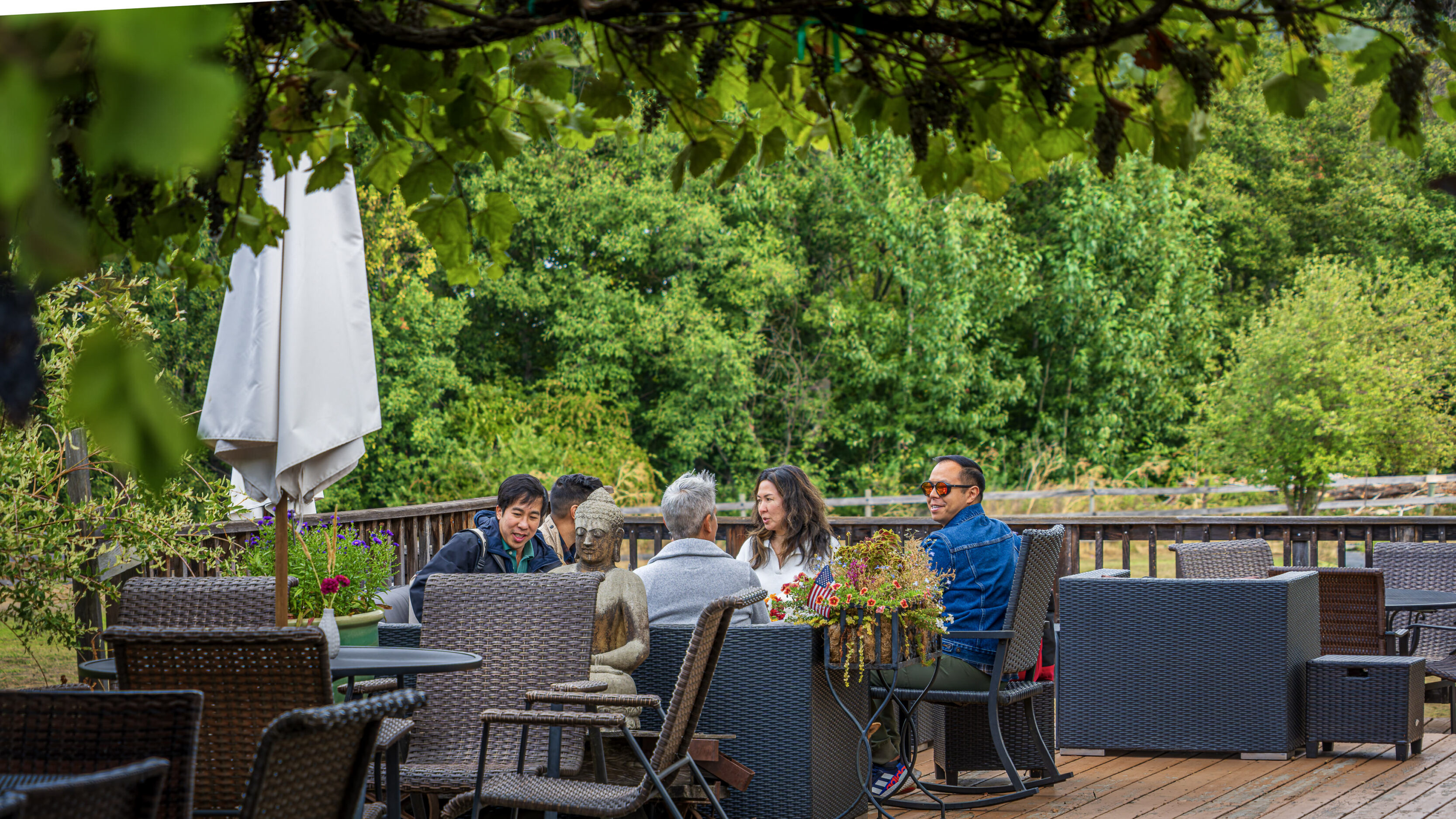 2026_08_29_Orchard_Winery_Scenic_0137-Edit_3840 ‣ In The Viewfinder Group of people seated at a wicker patio table outdoors, chatting with a Buddha statue centerpiece and lush greenery in the background.