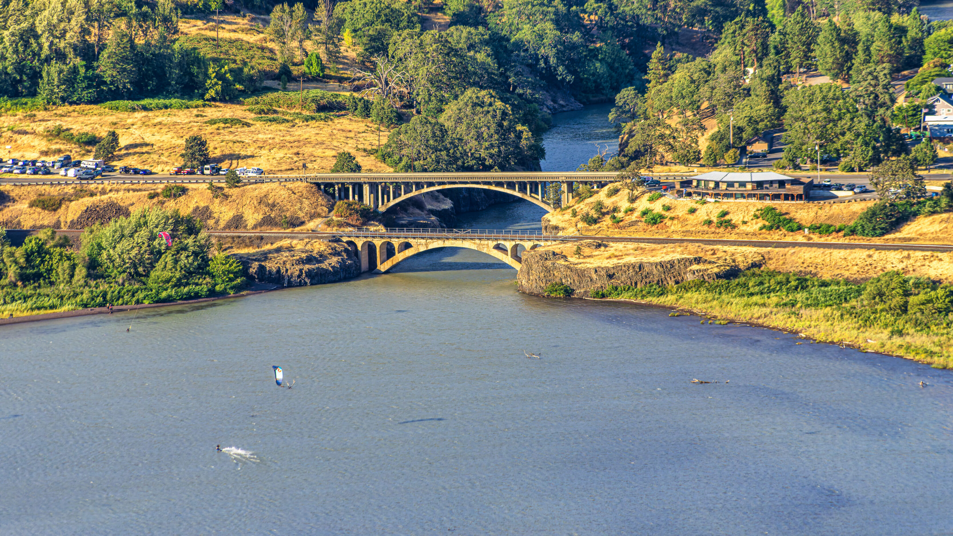2026_08_29_Orchard_Winery_Scenic_0299-Edit_3840 ‣ In The Viewfinder A river with two arched bridges crossing rocky banks, surrounded by green hills and trees, with a parking area and buildings along the shore.