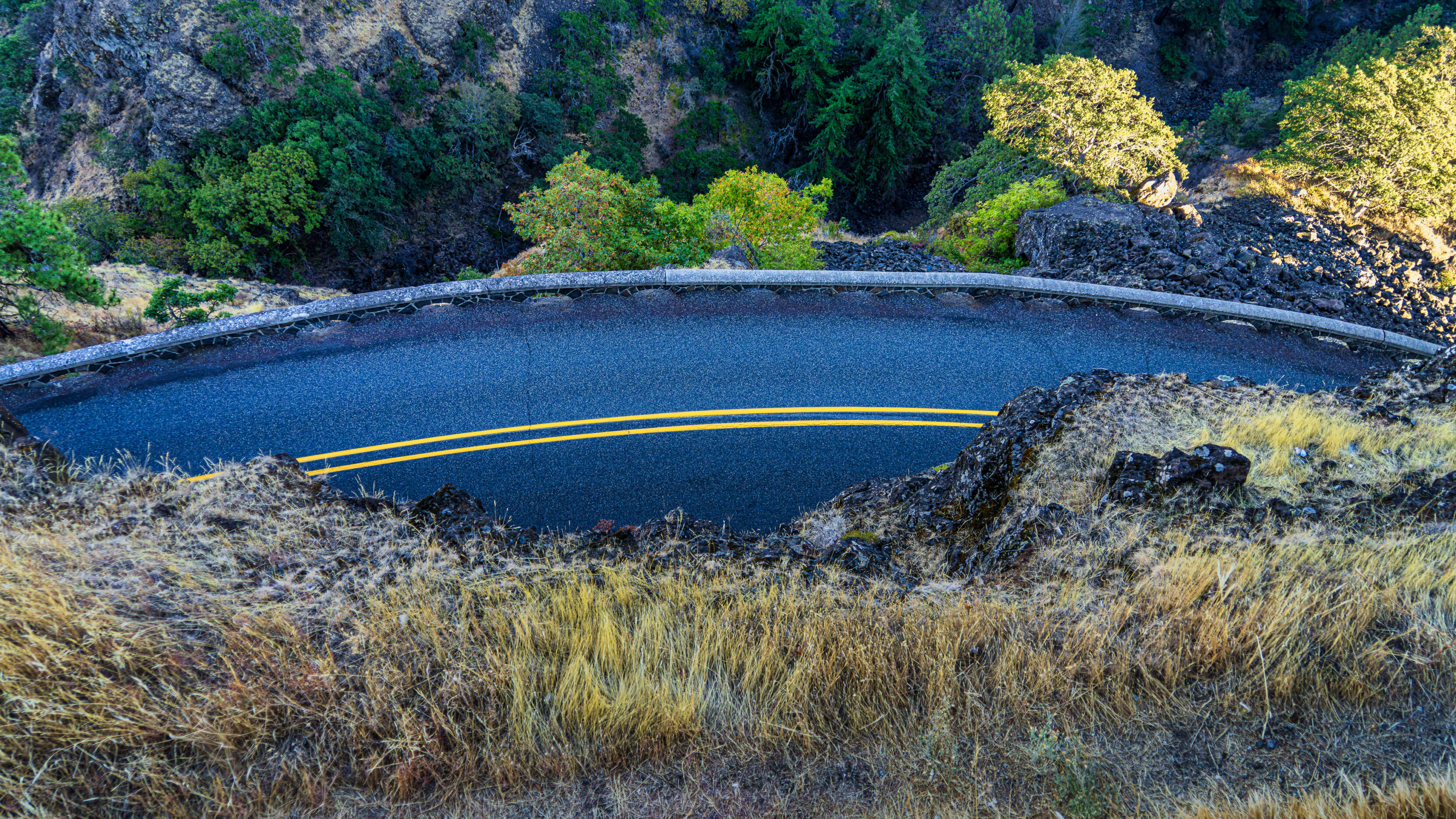 2026_08_29_Orchard_Winery_Scenic_0335-Edit_3840 ‣ In The Viewfinder Rowena Crest Viewpoint - A winding dark asphalt road with yellow center lines curves through a rocky hillside and sparse grass, flanked by a concrete guardrail and rugged terrain as trees overlook the road.