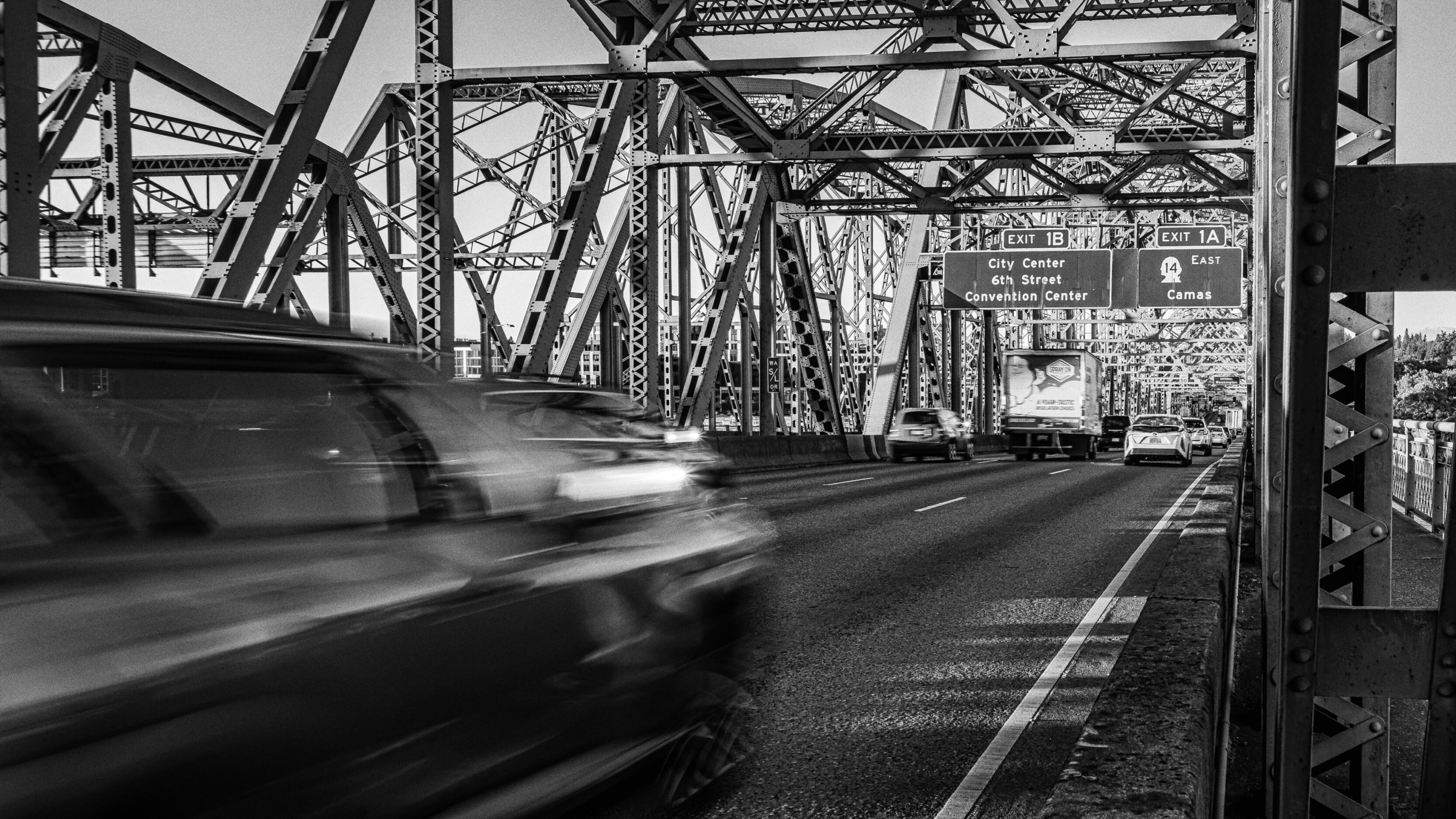 Black-and-white view of a steel truss bridge filled with moving traffic; exit signs and a highway lane guide the way toward the city center and convention center