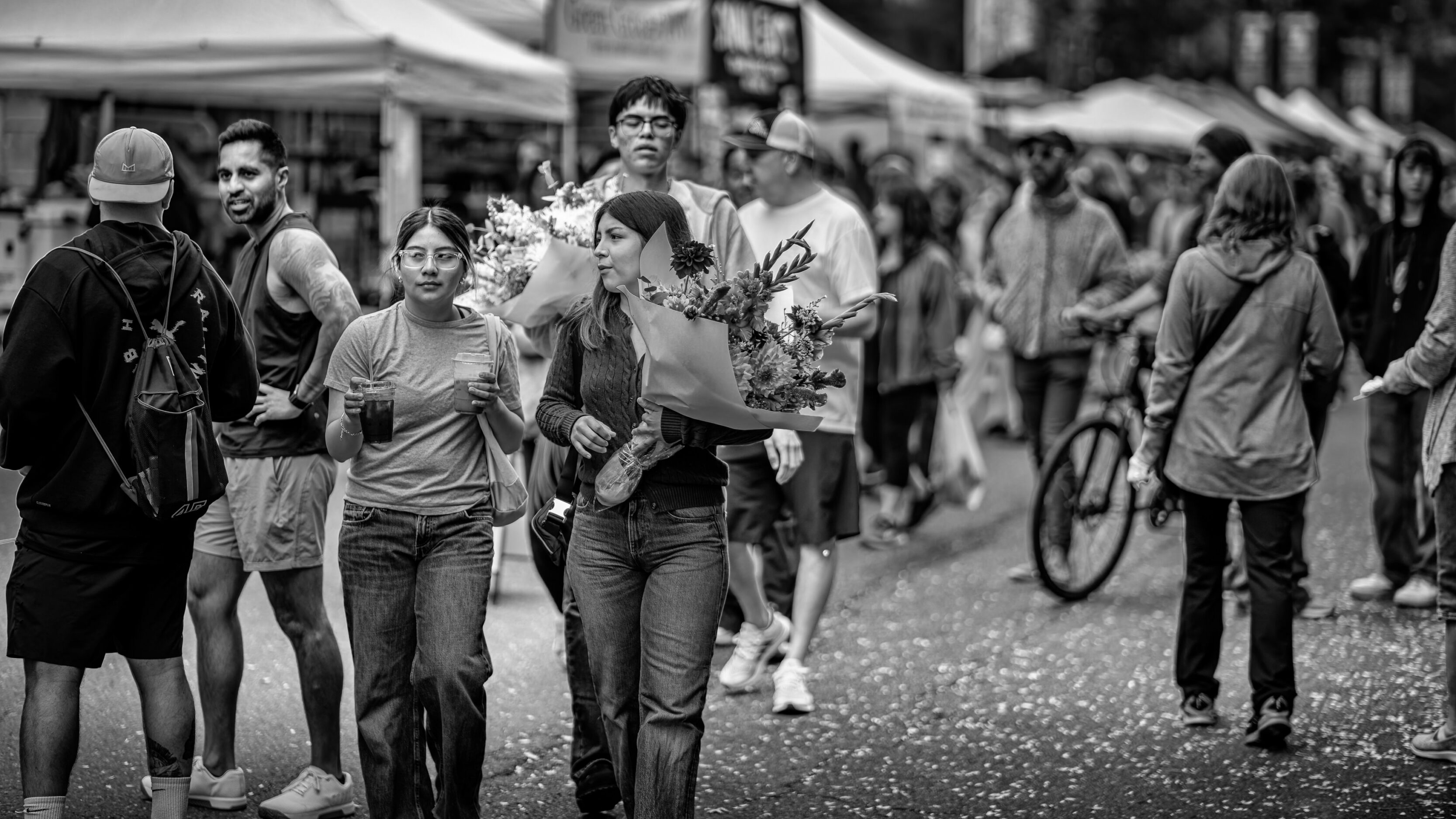 2026_09_19_Vancouver_85mm_0085-Edit-Edit_3840 ‣ In The Viewfinder People walk through an outdoor market; a woman in the center carries a bouquet wrapped in paper and others surround her.