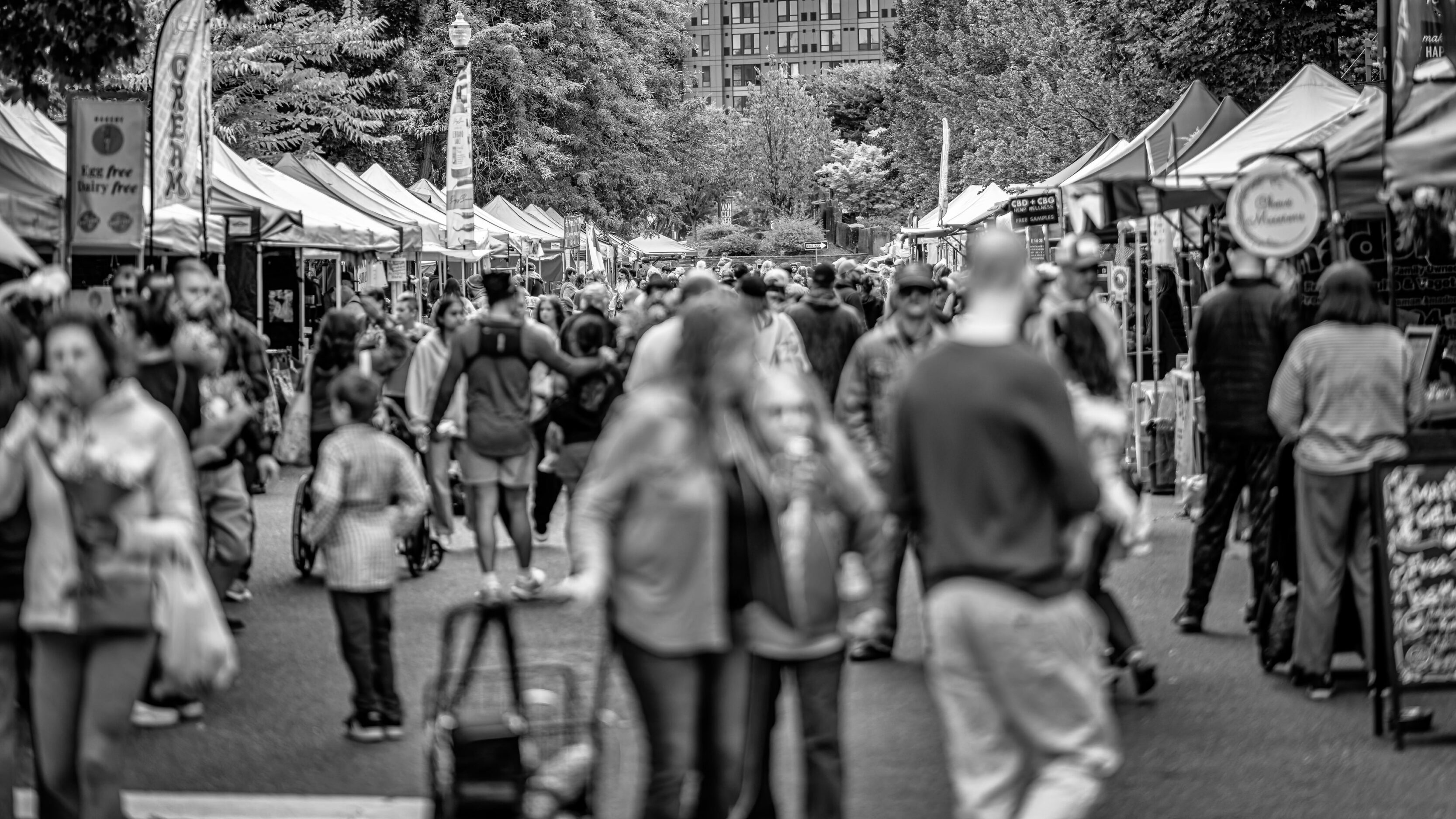 2026_09_19_Vancouver_85mm_0106-Edit-Edit_3840 ‣ In The Viewfinder Outdoor farmers market scene with stalls, shoppers, and a prominent banner reading'Welcome to the Market!' (Vancouver Farmers Market) in black and white.