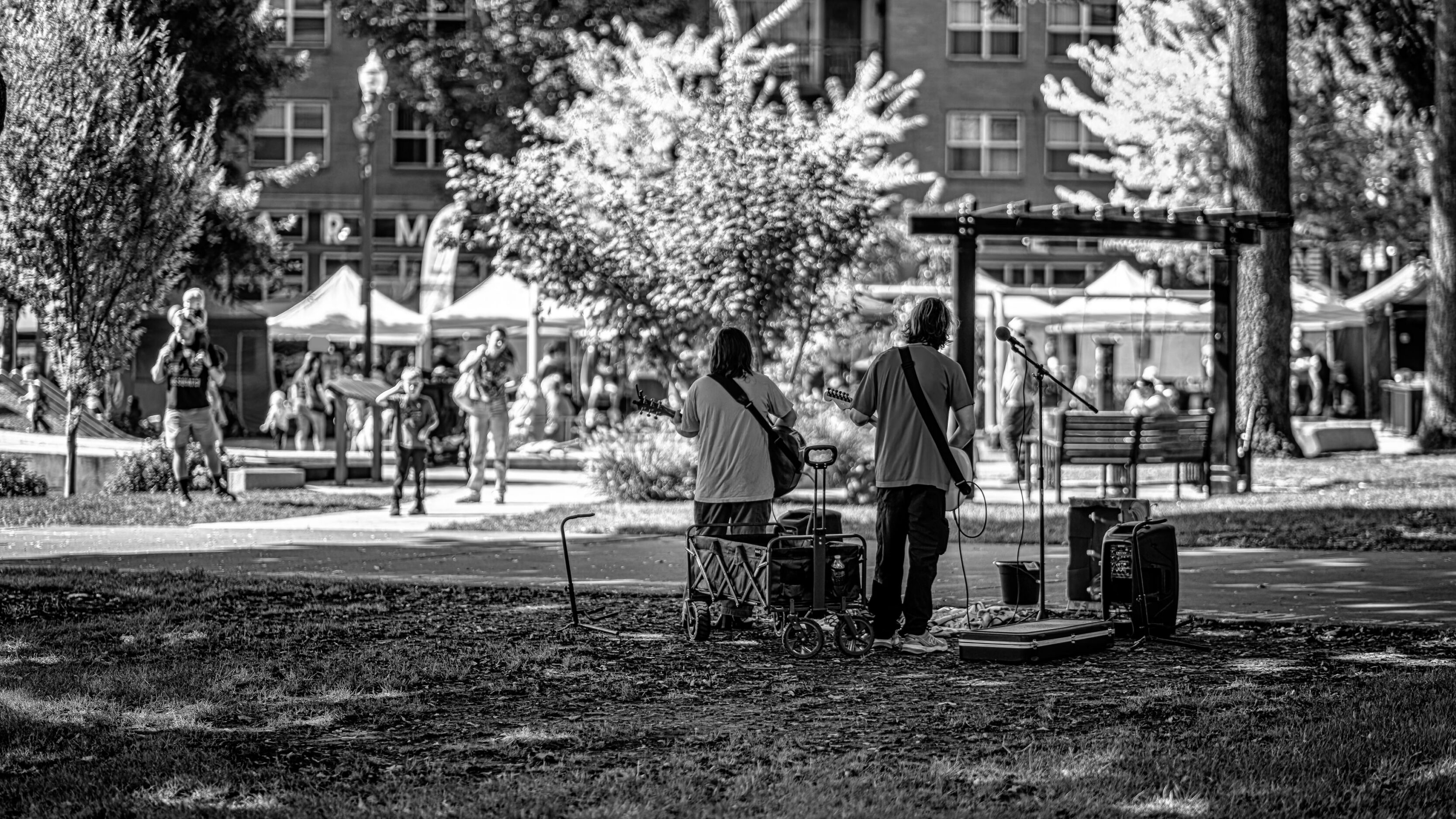 2026_09_19_Vancouver_85mm_0154-Edit-Edit_3840 ‣ In The Viewfinder Two guitarists perform outdoors in a park as a small stage setup and audience watch nearby stalls and tents nearby.