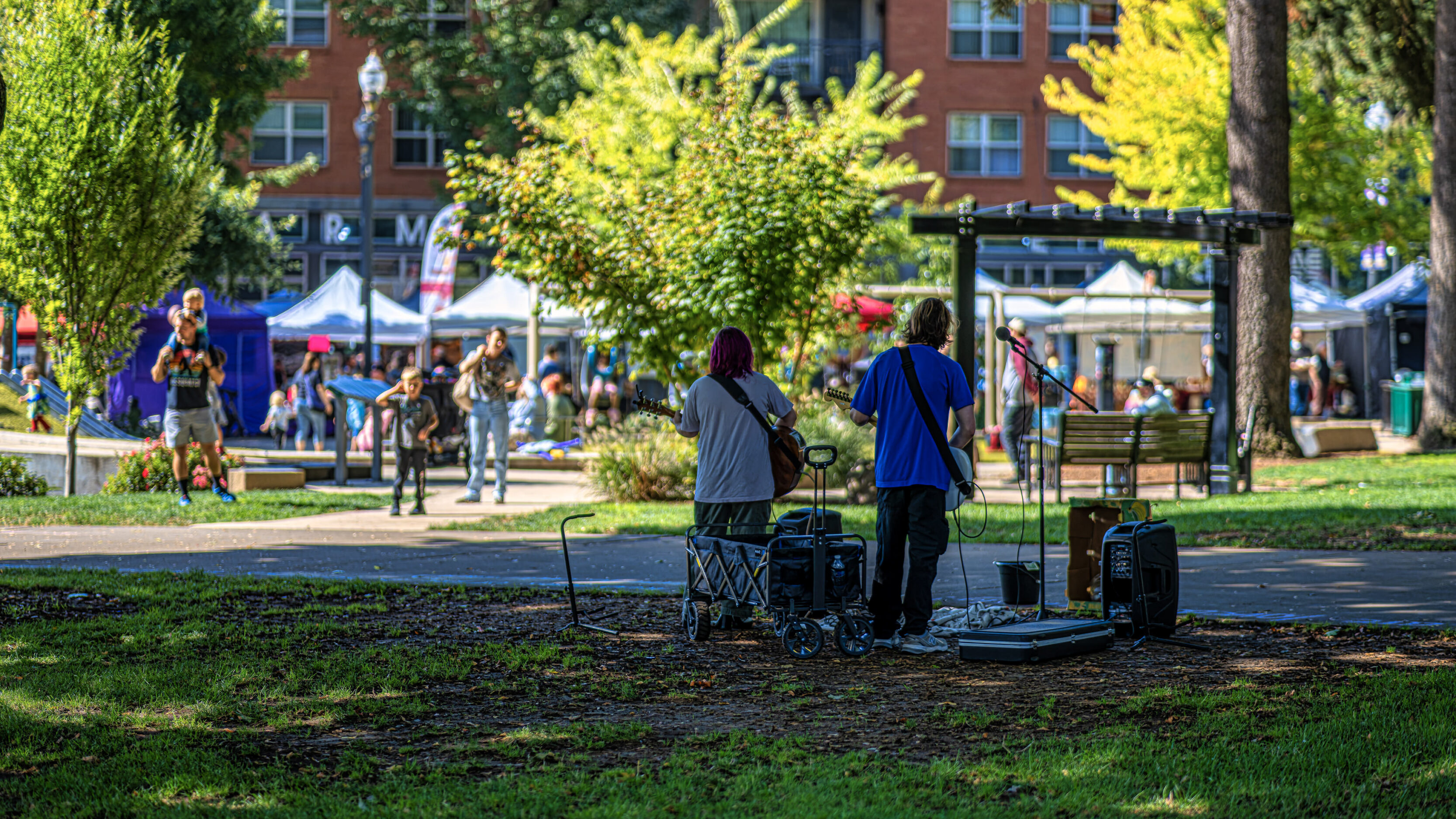 Two guitarists perform on a sunny park stage while a crowd and market tents appear in the background.