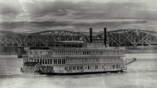 Historic paddlewheel steamboat on a wide river with a steel truss bridge in the background.