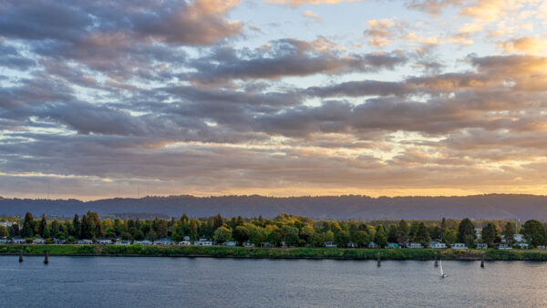 Sunset over a calm river, with a tree-lined shore and houses along the shoreline ahead.