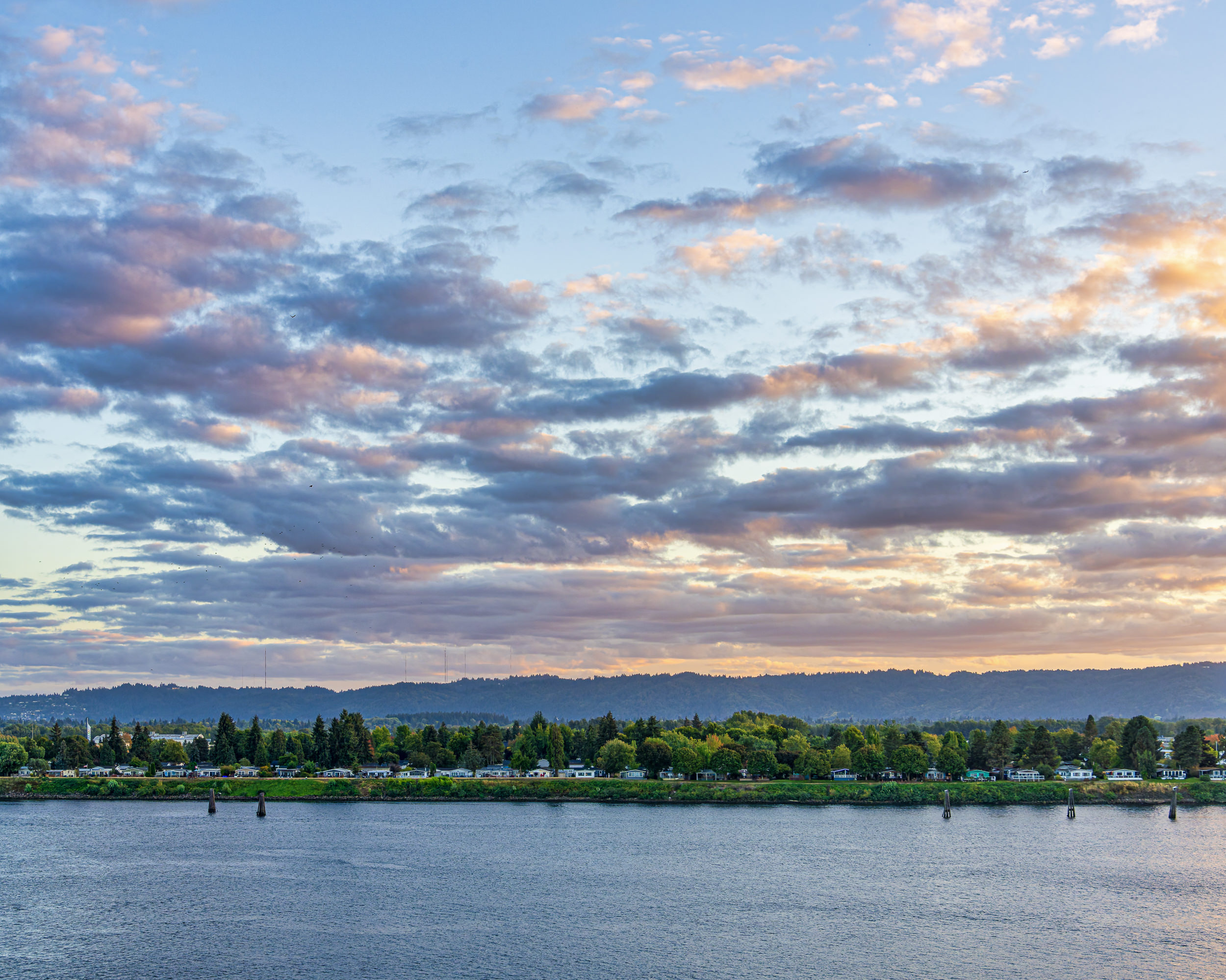 Lakeside scene at sunset with a tree-lined shore, houses along the water, and a sky with scattered clouds.