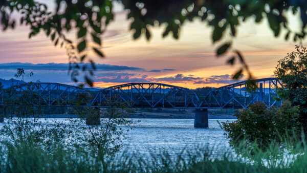 Sunset over a river with a metal truss bridge framed by foliage in the foreground and silhouettes of hills in the distance.