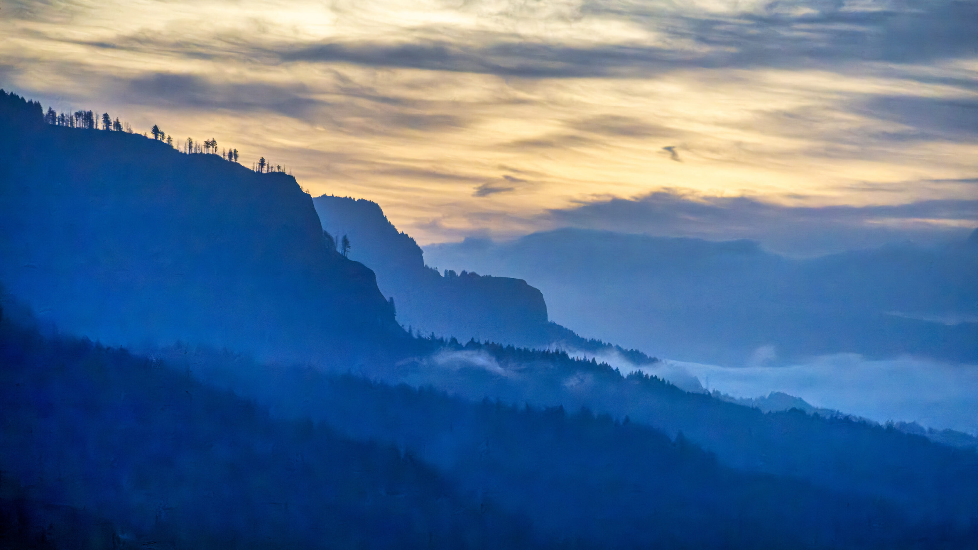 2026_09_26_Columbia_River_Gorge_0011-Edit_3840 ‣ In The Viewfinder Blue-toned mountain range fading into fog under a pale sunrise sky, silhouettes along the ridgeline.