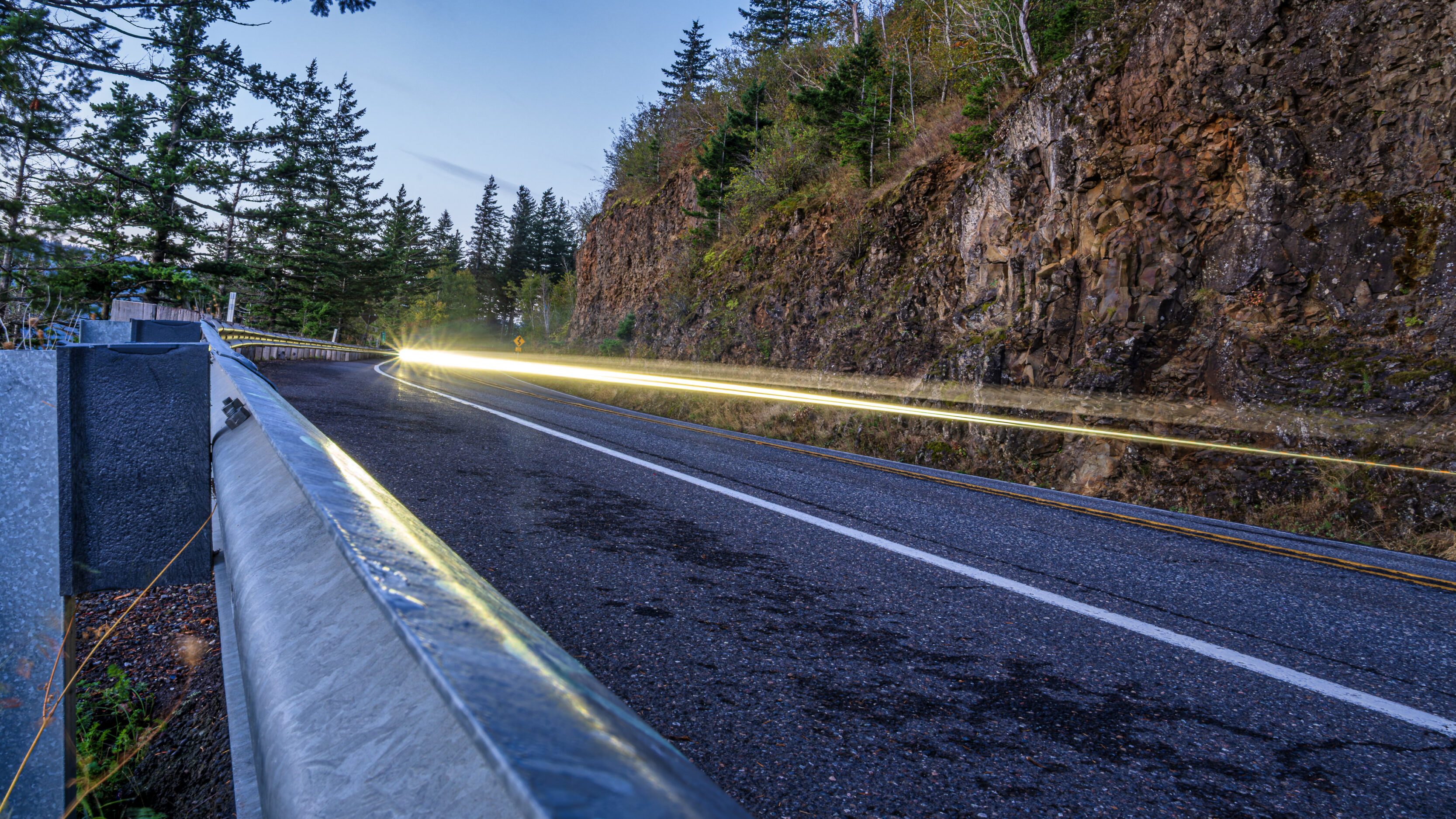 2026_09_26_Columbia_River_Gorge_0035-Edit_3840 ‣ In The Viewfinder Curved mountain road beside a rocky cliff, with a metal guardrail and evergreen forest on the left, light trails from passing cars.