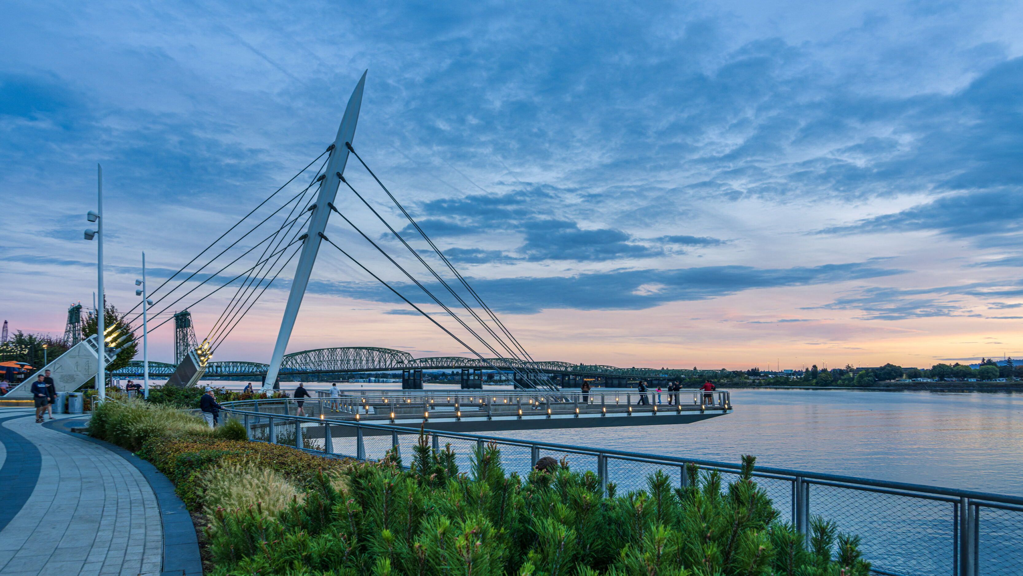 2026_09_28_Waterfront_Sunset_0024-Edit_3840 ‣ In The Viewfinder Public park by the water with a tall white angled cable-stayed sculpture, supporting cables, and a curved boardwalk at sunset; a bridge spans the river in the distance with people walking along the walkway towards the horizon.