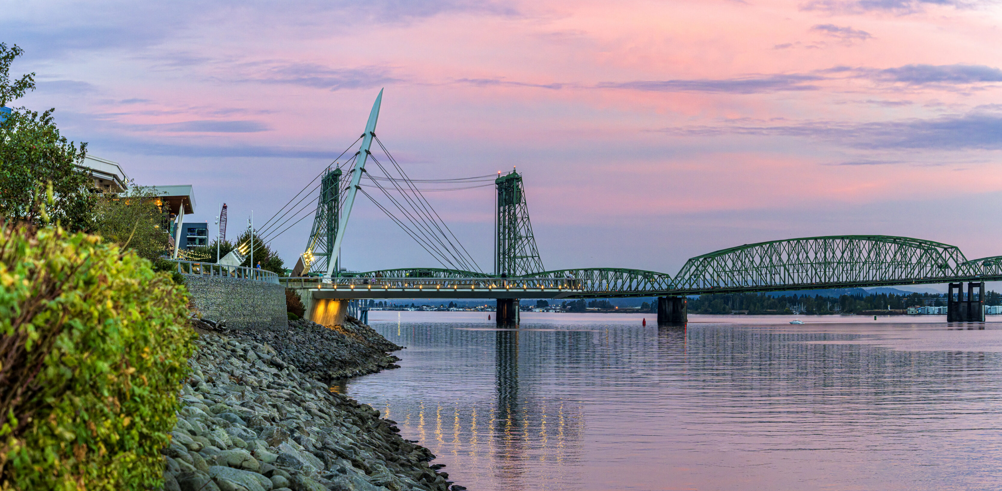 2026_09_28_Waterfront_Sunset_0038-Pano-Edit_3840 ‣ In The Viewfinder Green cable-stayed bridge over a calm river at sunset, with a rocky shoreline and pink/purple sky in the background.
