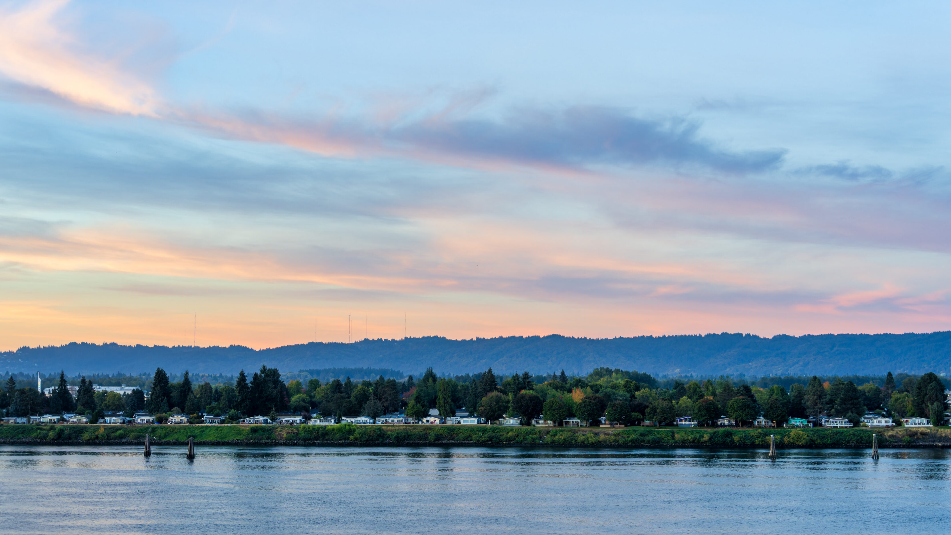 2026_10_01_Balcony_001-DxO_DeepPRIME 3-Edit_3840 ‣ In The Viewfinder River at dusk with calm water, tree-lined shoreline and a row of houses along the far bank under a pastel sunset sky with pink, blue, and orange hues, AM PM View From Our Balcony