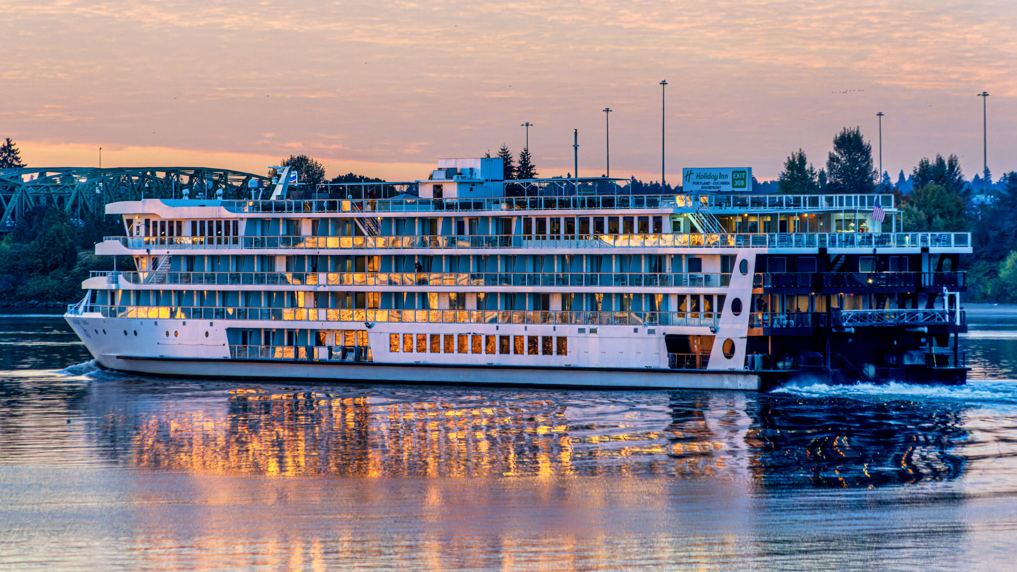 Sunset-lit multi-deck river cruise ship docked by the shore, its glowing windows reflecting on the water.