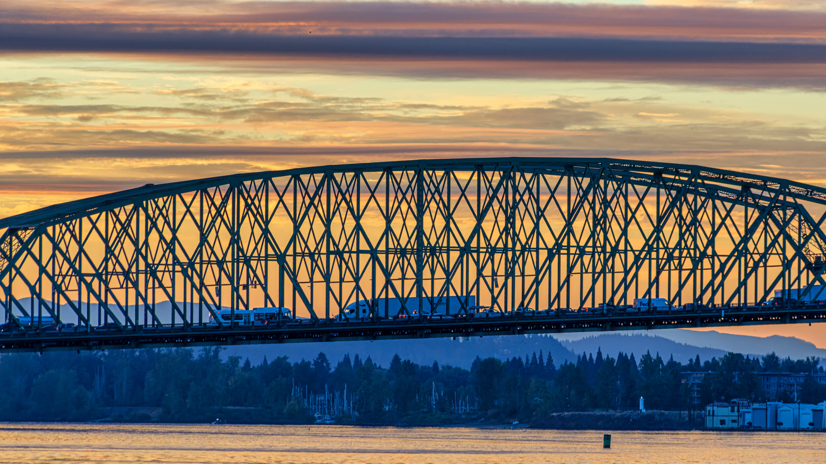 Blue steel lattice bridge spans a calm river at sunset, with a tree-lined shore and distant hills silhouetted below a colorful sky.