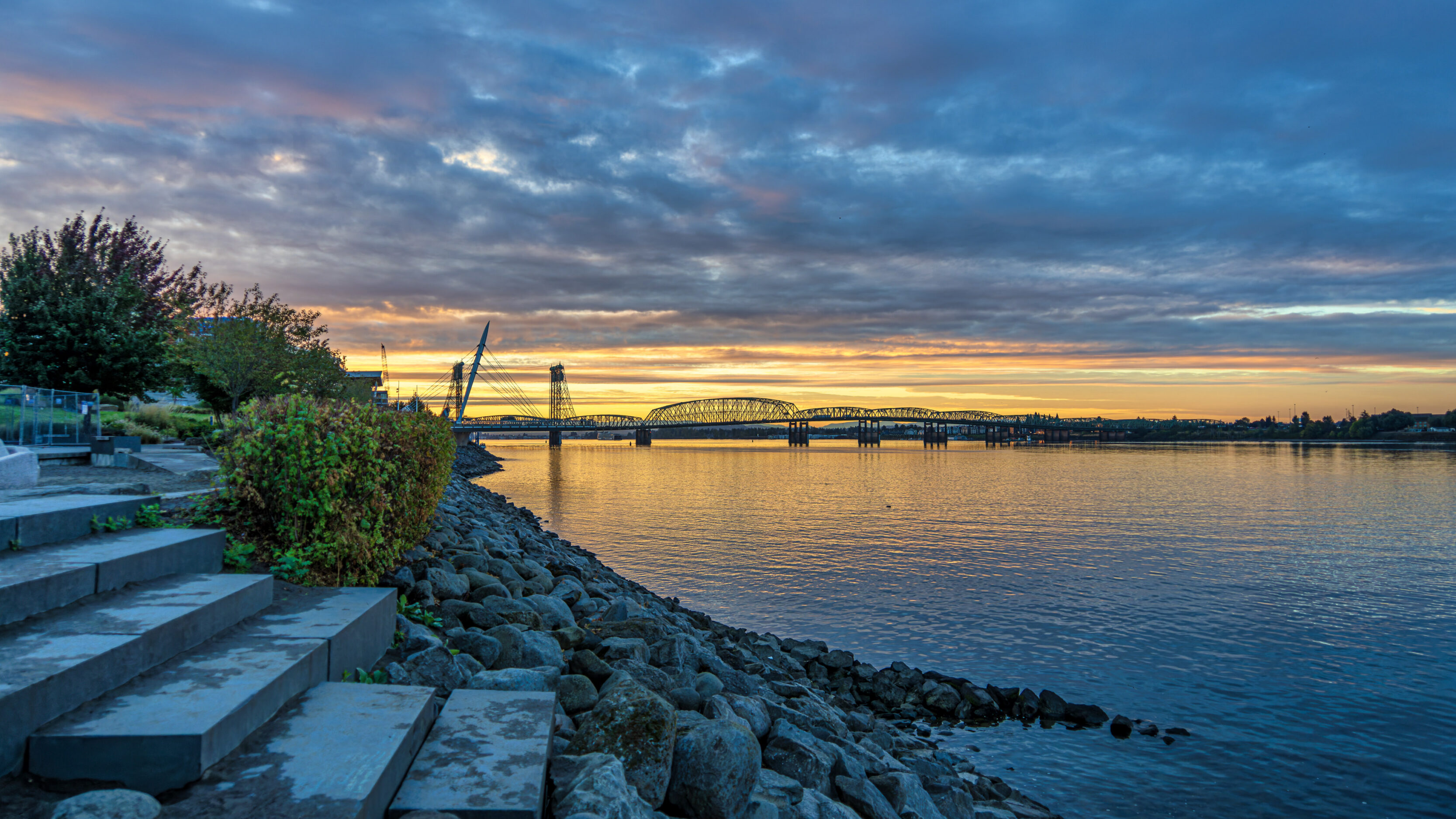 Rocks and concrete steps along a calm river at sunset, with a large bridge in the distance.