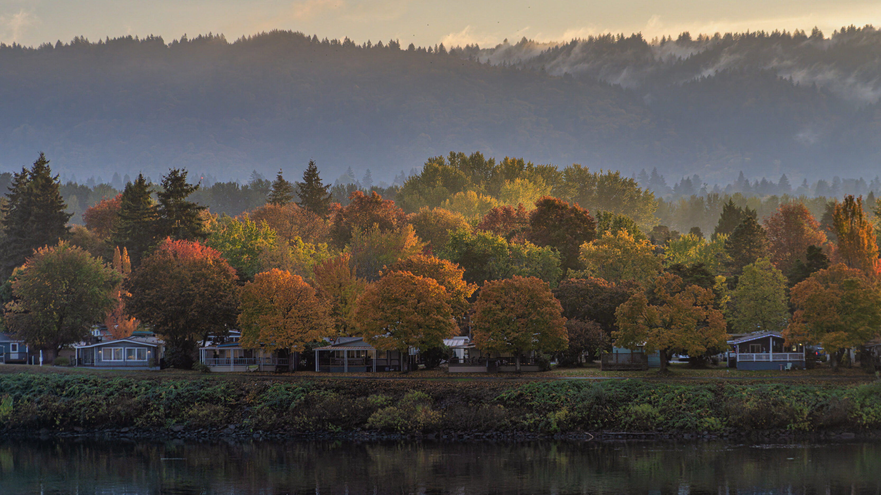 The View From Our Balcony Row of houses along a river with colorful autumn trees in front and misty forested hills beyond.