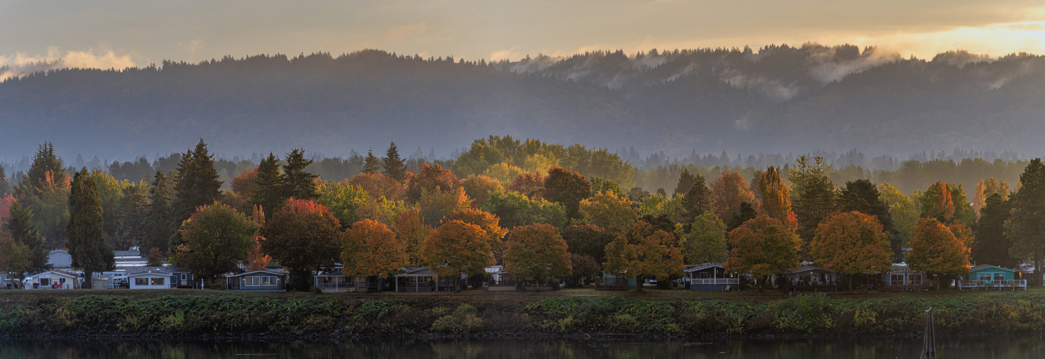 The View From Our Balcony Autumn trees line a lakeside with colorful foliage and small houses, misty hills in the background at dawn.